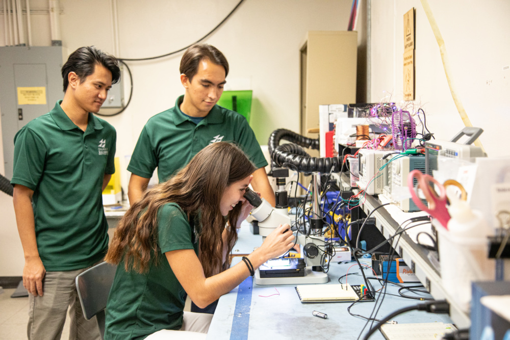 Three people in green shirts work in an electronics lab. One is using a microscope, while the other two observe. Equipment and tools are scattered on the workbench.